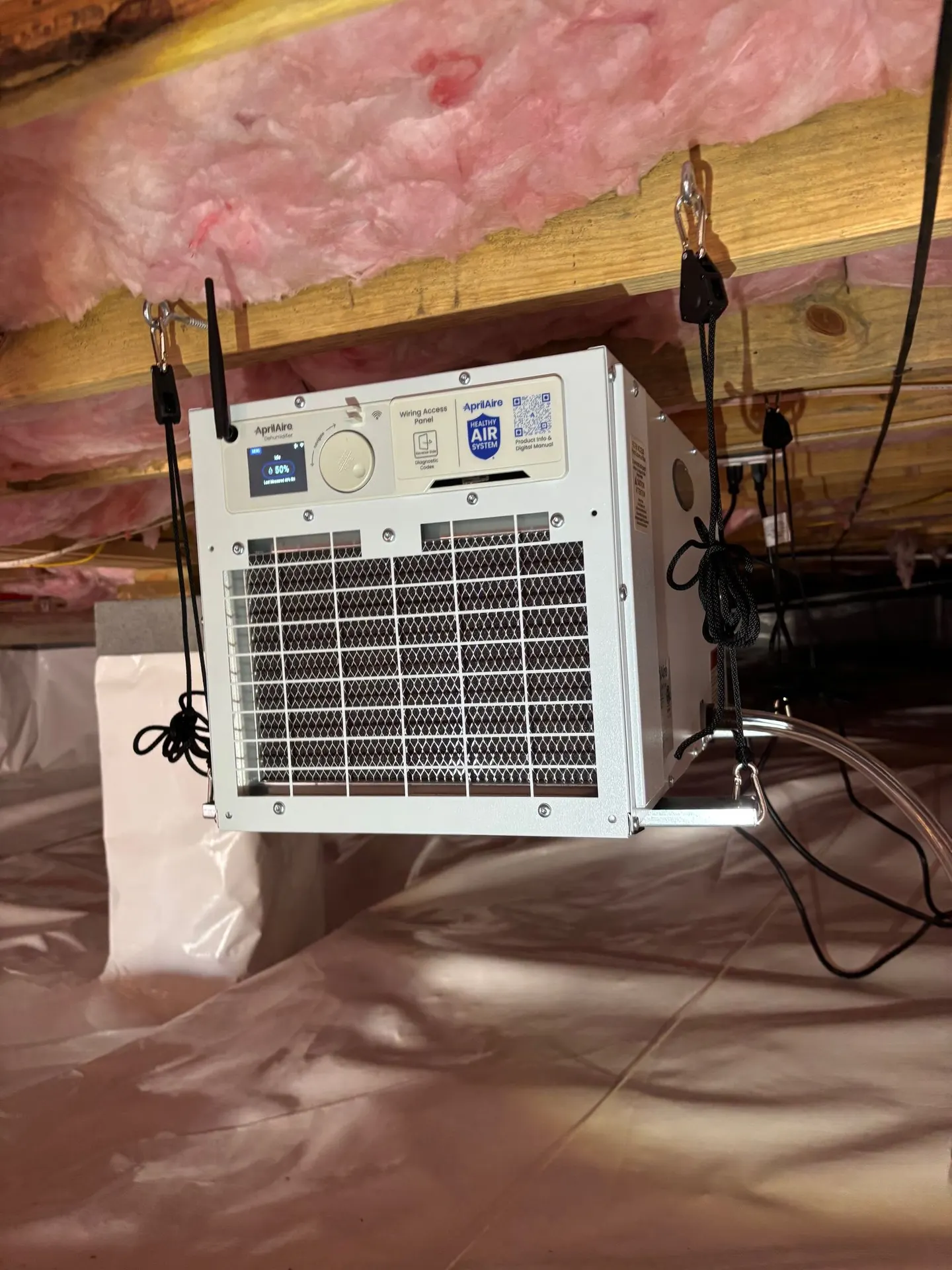 Dehumidifier hung from the floor joists of a crawl space, with a white vapor barrier covering the ground