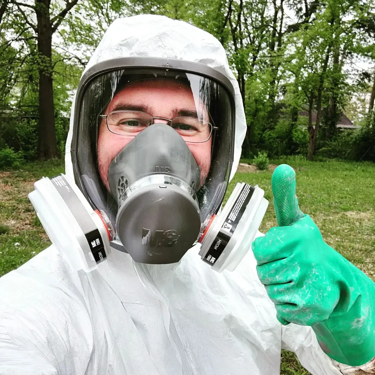 Trust Exterminating technician in a full-face respirator and white protective suit, giving a thumbs up outdoors